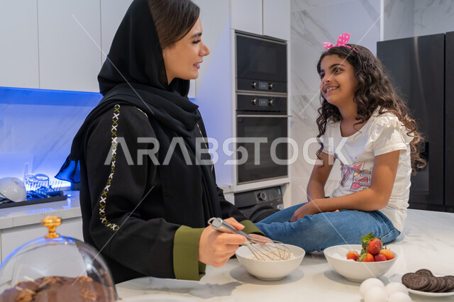 قضاء أوقات ممتعه مع الابنه في المطبخ، اعداد وتحضير الحلويات الطازجه في المنزل, الوقوف أمام طاولة المطبخ, مقادير صنع الكعك، ام عربيه خليجية امارتية محجبه تقف مع ابنتها يقومان بتحضير الطعام، عائلة سعودية سعيدة، المشاركة بالأعمال المنزلية، تعلم وصفات جديدة