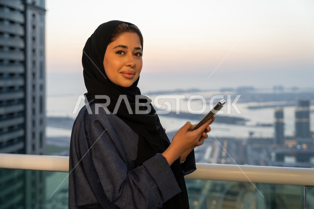 تصفح التطبيقات وبرامج التواصل الاجتماعي، تبادل الأحاديث والأخبار مع الأهل والأصدقاء، إمرأة عربية خليجية سعودية ترتدي العباءة والحجاب تمسك بيدها الهاتف المحمول، سيدة اماراتية تقوم باستخدام الأجهزة والتقنيات الحديثة والمتطورة، خلفية الابراج وناطحات السحاب