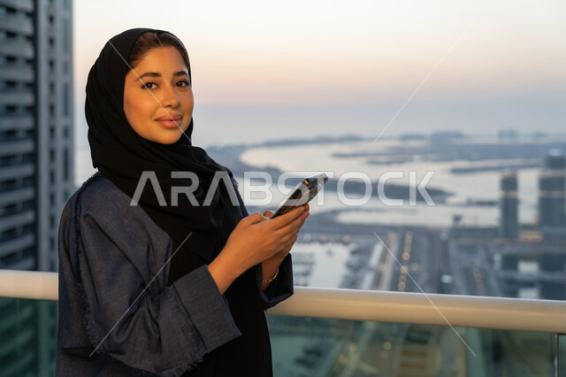 تصفح التطبيقات وبرامج التواصل الاجتماعي، تبادل الأحاديث والأخبار مع الأهل والأصدقاء، إمرأة عربية خليجية سعودية ترتدي العباءة والحجاب تمسك بيدها الهاتف المحمول، سيدة اماراتية تقوم باستخدام الأجهزة والتقنيات الحديثة والمتطورة، خلفية الابراج وناطحات السحاب