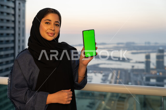 عرض شاشة خضراء فارغة كروما عبر الجوال، تصفح التطبيقات وبرامج التواصل الاجتماعي، إمرأة عربية خليجية سعودية ترتدي العباءة والحجاب تمسك بيدها الهاتف المحمول، سيدة اماراتية تقوم باستخدام الأجهزة والتقنيات الحديثة والمتطورة، خلفية الابراج وناطحات السحاب
