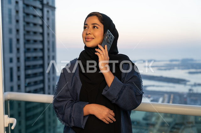 Communication and checking on friends and relatives, exchanging entertaining conversations via mobile phone, a Saudi Arabian Gulf woman wearing an abaya and hijab making a phone call, an Emirati woman using modern and advanced devices and technologies, the concept of kinship, a background of towers and skyscrapers