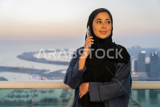 الاطمئنان على الأصدقاء والأقارب، تبادل الأحاديث المسليه عبر الجوال، إمرأة عربية خليجية سعودية ترتدي العباءة والحجاب تقوم باجراء مكالمة هاتفية، سيدة اماراتية تقوم باستخدام الأجهزة والتقنيات الحديثة والمتطورة، مفهوم صلة الرحم، خلفية الابراج وناطحات السحاب