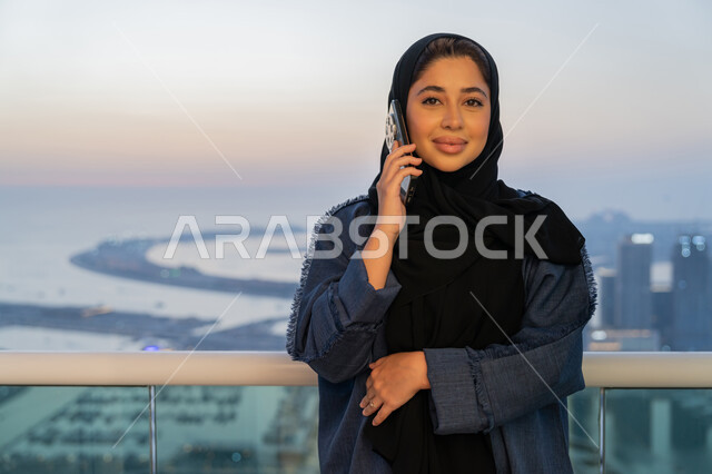 Communication and checking on friends and relatives, exchanging entertaining conversations via mobile phone, a Saudi Arabian Gulf woman wearing an abaya and hijab making a phone call, an Emirati woman using modern and advanced devices and technologies, the concept of kinship, a background of towers and skyscrapers