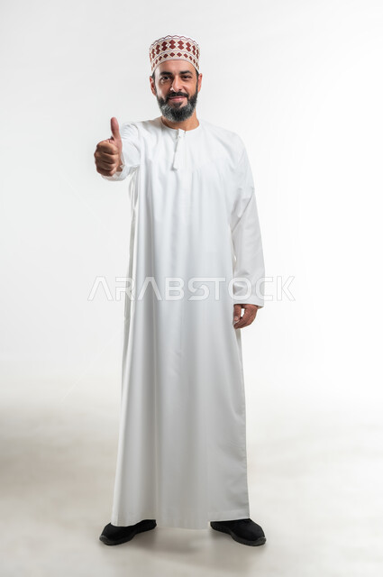ايماءات الرضا والسرور والسعادة، حركات الاعجاب والقبول, الموافقة على أمر معين، بورتريه لرجل عربي خليجي عماني مبتسم يرتدي الدشداشة و الكمه ينظر الى الكاميرا يرفع اصبعه الإبهام للأعلى بتعابير علامة الجودة, صورة الجسم بالطول الكامل، خلفية بيضاء