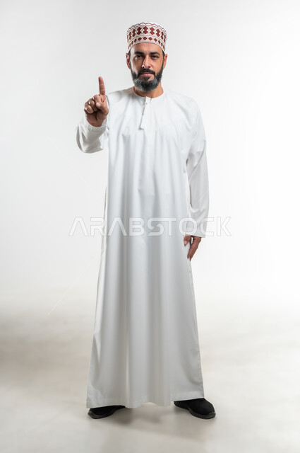 Accusation, reprimand and blame, request to stop doing wrong, feeling annoyed and wanting revenge, portrait of an Arab Gulf Omani man wearing a dishdasha and a sleeve raising his index finger forward to identify someone (you), full body image, white background