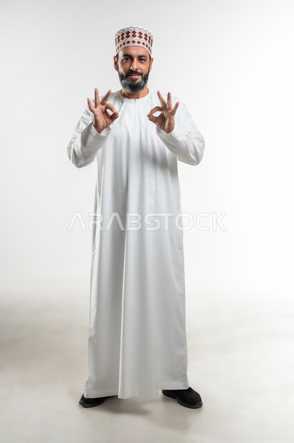 تعابير الرضا والسرور والسعادة، حركات الاعجاب والقبول, الموافقة على أمر معين، بورتريه لرجل عربي خليجي عماني مبتسم يرتدي الدشداشة و الكمه ينظر الى الكاميرا يرفع يديه للأعلى بإيماءات علامة الجودة, صورة الجسم بالطول الكامل، خلفية بيضاء