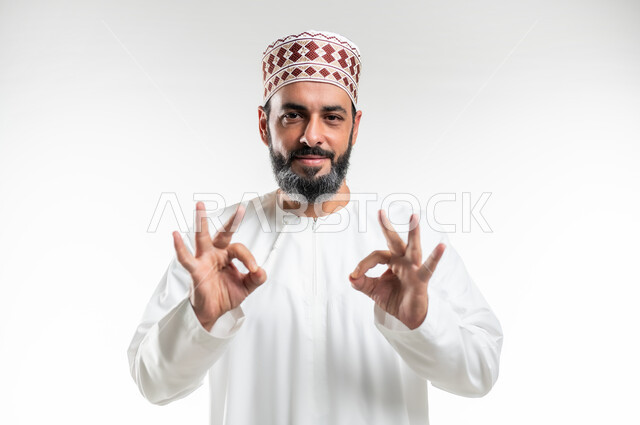 حركات الاعجاب والقبول, الموافقة على أمر معين، بورتريه مقرب لرجل عربي خليجي عماني مبتسم يرتدي الدشداشة و الكمه ينظر الى الكاميرا يرفع يديه للأعلى بإيماءات علامة الجودة, تعابير الرضا والسرور والسعادة، خلفية بيضاء