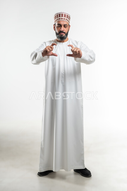 Using hands to measure something, determining volume and mass, portrait of an Arab Gulf Omani man wearing a dishdasha and a kummah standing straight and raising his hands in gestures of carrying something, concept of presenting and displaying a certain item, full-length body image, white background