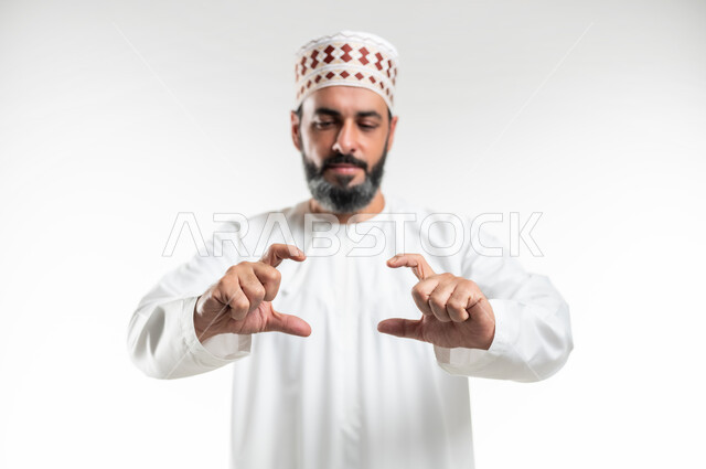 استخدام اليدين لتحجيم شيء ما، تحديد الحجم والكتله، بورتريه مقرب لرجل عربي خليجي عماني يرتدي الدشداشة و الكمه يقف باستقامة ويقوم برفع يديه بايماءات حمل شيء ما، مفهوم تقديم وعرض عنصر معين، خلفية بيضاء