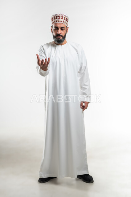 إحتواء وإمساك عنصر معين، حركات وإشارات يدين تدل على رفع الأشياء، بورتريه لرجل عربي خليجي عماني يرتدي الدشداشة العمانية والكمه يقف باستقامة ويرفع يده الى الأعلى بتعابير حمل شيء ما، صورة الجسم بالطول الكامل، خلفية بيضاء
