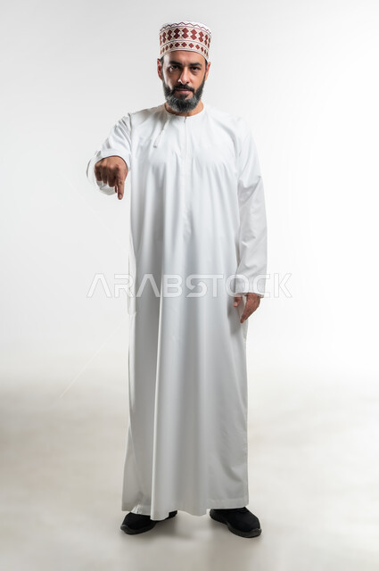 Pointing to a specific location, expressions of specificity and emphasis, signs that identify this place (here), portrait of an Arab Gulf Omani man wearing a dishdasha and a sleeve standing straight and pointing his index fingers down, full-length body image, white background