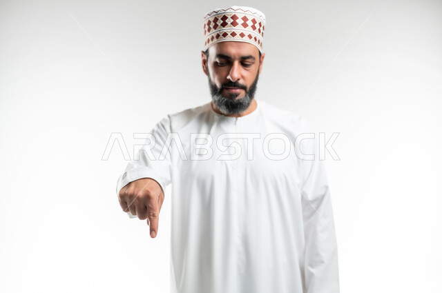 اشارات تحدد هذا المكان (هنا)، الاشارة إلى موقع معين، تعابير التحديد والتأكيد, بورتريه مقرب لرجل عربي خليجي عماني يرتدي الدشداشة والكمه يقف باستقامة و يشير بأصابع السبابة للأسفل ، خلفية بيضاء