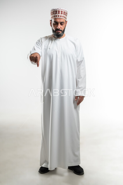 تعابير التحديد والتأكيد, اشارات تحدد هذا المكان (هنا)، الاشارة إلى موقع معين، بورتريه لرجل عربي خليجي عماني يرتدي الدشداشة والكمه يقف باستقامة و يشير بأصابع السبابة للأسفل ، صورة الجسم بالطول الكامل، خلفية بيضاء
