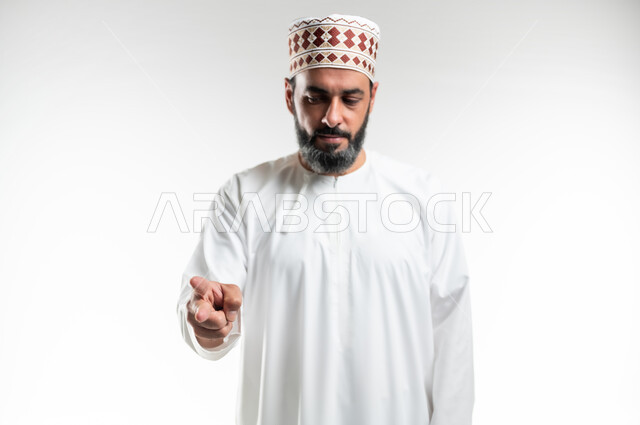 Pointing to something, gestures to specify this place (here), expressions of specification and emphasis, close-up portrait of an Arab Gulf Omani man wearing a dishdasha and a kemeh standing straight and pointing with two fingers to choose, white background