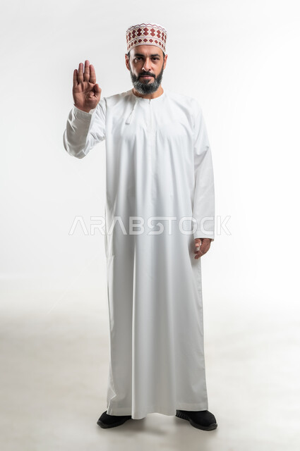 ايماءات الحزم والجدية, طلب التوقف وعدم الاقتراب, مفهوم الرفض والامتناع والانزعاج, بورتريه لرجل عربي خليجي عماني يرتدي الدشداشة والكمه يمد يده نحو الأمام مع رفع الكف بتعابير النهي عن فعل امر ما, صورة الجسم بالطول الكامل، خلفية بيضاء