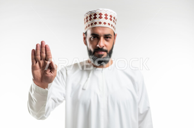 مفهوم الرفض والامتناع والانزعاج, ايماءات الحزم والجدية, طلب التوقف وعدم الاقتراب, بورتريه مقرب لرجل عربي خليجي عماني يرتدي الدشداشة والكمه يمد يده نحو الأمام مع رفع الكف بتعابير النهي عن فعل امر ما, خلفية بيضاء