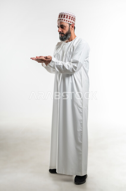 مفهوم الطلب والحاجة إلى مساعدة، تقديم وعرض شيء ما، بورتريه لرجل عربي خليجي عماني مبتسم يرتدي الدشداشة و الكمه يرفع يديه للأعلى مع فتح الكفين بإيماءات محاولة حمل عنصر ما، صورة الجسم بالطول الكامل، خلفية بيضاء
