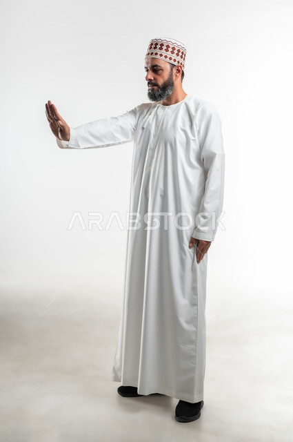 تعابير الحزم والجدية, طلب التوقف وعدم الاقتراب, مفهوم الرفض والامتناع والانزعاج, بورتريه لرجل عربي خليجي عماني يرتدي الدشداشة والكمه يمد يده نحو الأمام مع رفع الكف بإيماءات النهي عن فعل امر ما, صورة الجسم بالطول الكامل، خلفية بيضاء