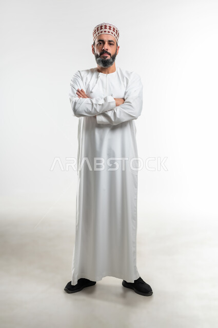الاهتمام بالمظهر الخارجي، الوقوف بإستقامة وأيدي متقاطعة، مفهوم الأناقة والرجولة، بورتريه لرجل عربي خليجي عماني مبتسم يرتدي الدشداشة والكمه ينظر إلى الكاميرا بإيماءات الثقة بالنفس، صورة الجسم بالطول الكامل، خلفية بيضاء