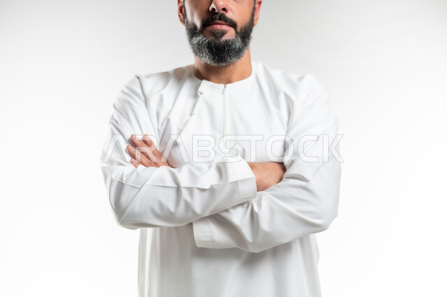 مفهوم الأناقة والرجولة، الاهتمام بالمظهر الخارجي، الوقوف بإستقامة وأيدي متقاطعة، بورتريه مقرب لرجل عربي خليجي عماني مبتسم يرتدي الدشداشة ينظر إلى الكاميرا بإيماءات الثقة بالنفس، خلفية بيضاء
