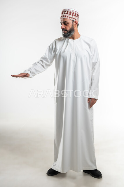 تعابير واشارات يدين تدل على قياس مسافة ما، تحديد طول وإرتفاع شيء ما، بورتريه لرجل عربي خليجي عماني يرتدي الكمه والدشداشة التقليدية العمانية يمد راحة كفه نحو الأسفل بايماءات وجود عنصر معين، صورة الجسم بالطول الكامل، خلفية بيضاء