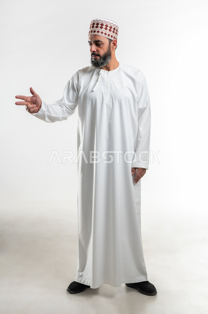 Gestures to identify this place (here), expressions of identification and emphasis, pointing to something, portrait of an Arab Gulf Omani man wearing a dishdasha and a sleeve standing straight and pointing with three fingers to choose, full-length body image, white background