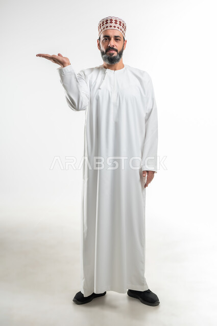 الوقوف بإستقامة والنظر الى الكاميرا، عرض عنصر محدد، بورتريه لرجل عربي خليجي عماني يرتدي الدشداشة و الكمة يرفع يده للأعلى لتقديم شيء ما، إيماءات وتعابير يدين تدل على حمل ورفع الأشياء، صورة الجسم بالطول الكامل، خلفية بيضاء