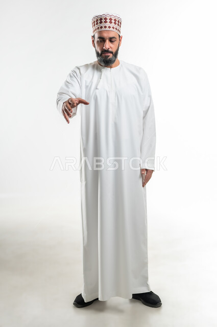 مفهوم الترحيب و السلام و إلقاء التحية، محاولة الإمساك بشيء ما، بورتريه لرجل عربي خليجي عماني يرتدي الدشداشة و الكمة يقوم بمد يده نحو الأمام لإلتقاط عنصر ما، حركات يدين تدل على المصافحة، الطول الكامل للجسم، خلفية بيضاء