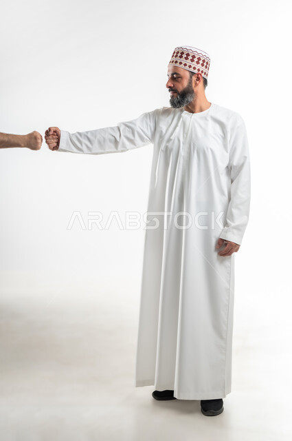 بناء علاقات و تكوين صداقات جديدة، الترحيب و السلام و إلقاء التحية، بورتريه لرجل عربي خليجي عماني يرتدي الدشداشة و الكمه يقوم توجيه قبضة يده مقابل يد شخص آخر، ايماءات وحركات يدين تدل على التشجيع، صورة الجسم بالطول الكامل، خلفية بيضاء