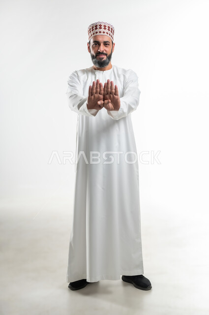 Warning against doing something, request to stop and not approach, concept of refusal, abstention and annoyance, portrait of an Arab Gulf Omani man wearing a dishdasha and a sleeve, smiling, extending his hands forward in gestures of forbidding doing something, full body length, white background
