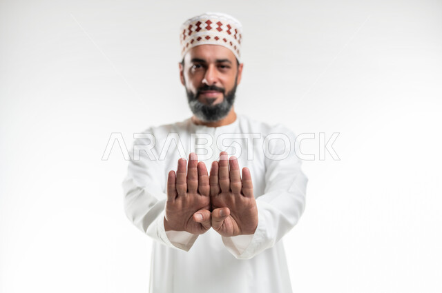 طلب التوقف وعدم الاقتراب, مفهوم الرفض والامتناع والانزعاج, بورتريه مقرب لرجل عربي خليجي عماني يرتدي الدشداشة والكمه مبتسم يمد كفيه نحو الأمام بإيماءات النهي عن فعل امر ما, التحذير من فعل شيء ما، خلفية بيضاء