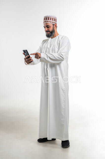 المحادثات مع الأهل والأصدقاء، إجراء مكالمات الفيديو عبر الهاتف المحمول، مشاهدة الأفلام والمسلسلات الممتعة، استخدام التكنولوجيا و التقنيات الحديثة, بورتريه لرجل عربي خليجي عماني يرتدي الدشداشة والكمه يقوم بالنقر على الموبايل، الطول الكامل للجسم، خلفية بيضاء