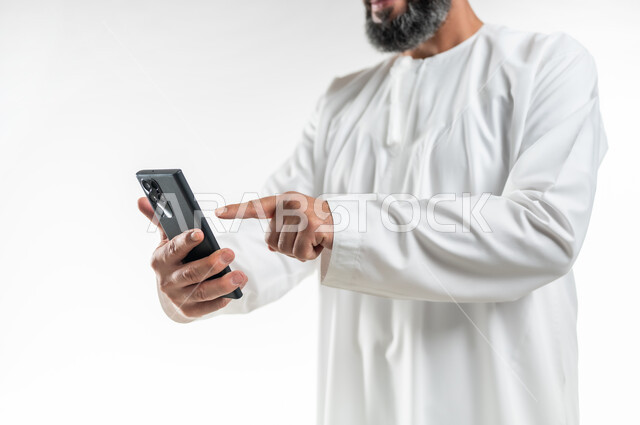 الدردشات والمحادثات مع الأهل والأصدقاء، إجراء مكالمات الفيديو عبر الهاتف المحمول، مشاهدة الأفلام والمسلسلات الممتعة، استخدام التكنولوجيا و التقنيات الحديثة, بورتريه مقرب لرجل عربي خليجي عماني يرتدي الدشداشة يقوم بالنقر على الموبايل، خلفية بيضاء