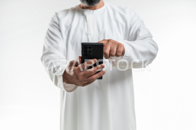 التواصل مع الأهل والأصدقاء، إجراء مكالمات الفيديو عبر الهاتف المحمول، مشاهدة الأفلام والمسلسلات الممتعة، استخدام التكنولوجيا و التقنيات الحديثة, بورتريه مقرب لرجل عربي خليجي عماني يرتدي الدشداشة يقوم بالنقر على الموبايل، خلفية بيضاء
