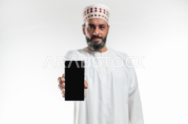 Mobile applications and services display, digital and electronic marketing concept, close-up portrait of an Arab Gulf Omani man wearing a dishdasha and a turban holding a mobile phone with a blank black screen, using advanced technology, white background
