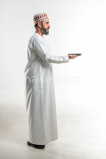 مفهوم التسويق الرقمي و الالكتروني، عرض تطبيقات وخدمات الهاتف المحمول، بورتريه لرجل عربي خليجي عماني يرتدي الدشداشة والكمة يحمل بيده موبايل حديث، استخدام التكنولوجيا الحديثة والتقنيات المتطورة، صورة الجسم بالطول الكامل، خلفية بيضاء