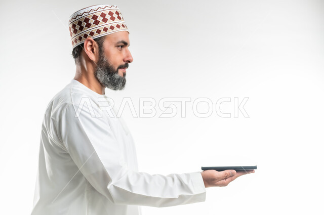 استخدام التكنولوجيا الحديثة والتقنيات المتطورة، مفهوم التسويق الرقمي و الالكتروني، عرض تطبيقات وخدمات الهاتف المحمول، بورتريه مقرب لرجل عربي خليجي عماني يرتدي الدشداشة والكمة يحمل بيده موبايل حديث، خلفية بيضاء