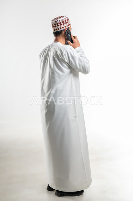 دردشات ومحادثات عبر الانترنت، إجراء المكالمات الصوتية، بورتريه من الخلف لرجل عربي خليجي عماني يرتدي الدشداشة يستخدم الهاتف المحمول، التواصل مع الأصدقاء عبر مواقع التواصل الاجتماعي، استخدام التكنولوجيا و التقنيات الحديثة, الطول الكامل للجسم، خلفية بيضاء