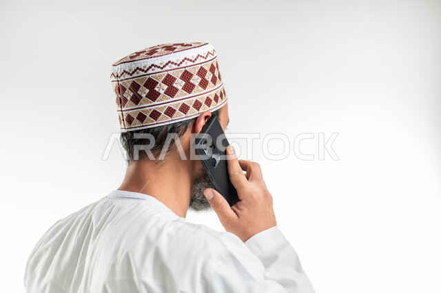 إجراء المكالمات الصوتية، دردشات ومحادثات عبر الانترنت، بورتريه مقرب من الخلف لرجل عربي خليجي عماني يرتدي الدشداشة يستخدم الهاتف المحمول، التواصل مع الأصدقاء عبر مواقع التواصل الاجتماعي، استخدام التكنولوجيا و التقنيات الحديثة, خلفية بيضاء