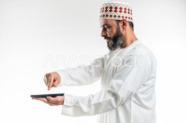 تصفح مواقع التواصل الاجتماعي عبر الهاتف المحمول، مشاهدة الأفلام والمسلسلات الممتعة، التواصل مع الأهل والأصدقاء، استخدام التكنولوجيا و التقنيات الحديثة, بورتريه مقرب لرجل عربي خليجي عماني يرتدي الدشداشة يقوم بالنقر على الموبايل، خلفية بيضاء