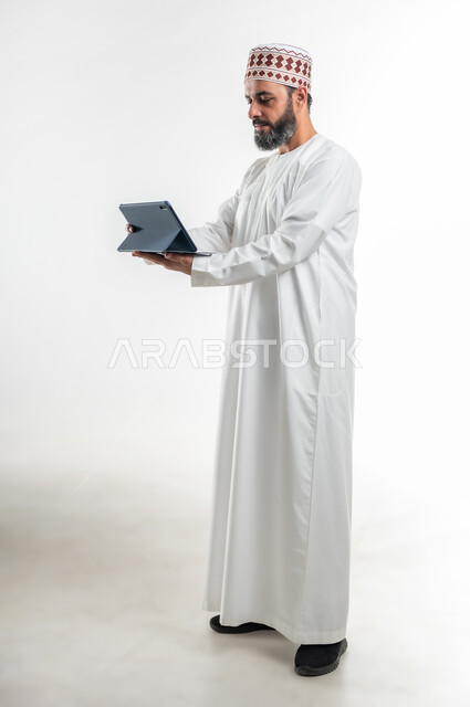 تصفح مواقع التواصل الاجتماعي، متابعة الأفلام والمسلسلات الممتعة، المحادثة مع الأهل والأصدقاء، استعمال التكنولوجيا و التقنيات الحديثة, بورتريه لرجل عربي خليجي عماني يرتدي الدشداشة والكمة يحمل بيده جهاز التابلت، الطول الكامل للجسم، خلفية بيضاء
