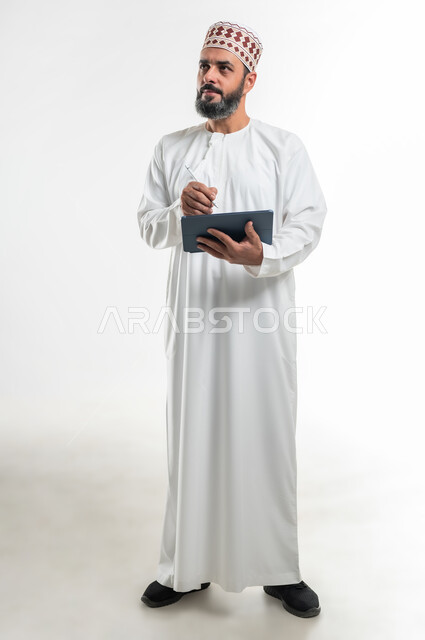 برامج وتطبيقات الأجهزة اللوحية، المحادثة مع الأهل والأصدقاء، استعمال التكنولوجيا و التقنيات الحديثة, بورتريه لرجل عربي خليجي عماني يرتدي الدشداشة والكمة يقوم بالكتابة على جهاز التابلت، تصفح برامج التواصل الاجتماعي، الطول الكامل للجسم، خلفية بيضاء