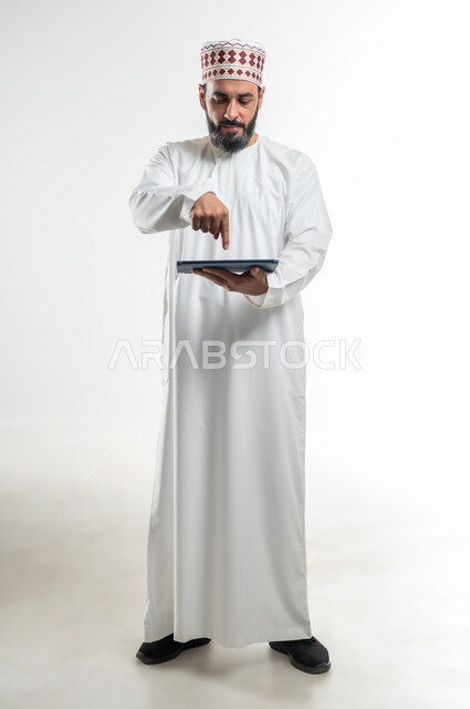 الدردشة مع الأهل والأصدقاء، استخدام التكنولوجيا و التقنيات الحديثة , بورتريه لرجل عربي خليجي عماني يرتدي الدشداشة والكمة يقوم بالنقر على جهاز التابلت، تصفح برامج التواصل الاجتماعي، تطبيقات وبرامج الأجهزة اللوحية، صورة الجسم بالطول الكامل , خلفية بيضاء