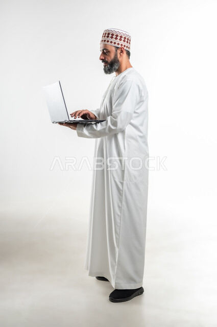 الاجتهاد وانجاز الاعمال المطلوبة، متابعة الأفلام والمسلسلات، تصفح مواقع التواصل الإجتماعي، بورتريه جانبي لرجل عربي خليجي عماني يرتدي الدشداشة والكمه يقف ويمسك بيده جهاز الحاسوب المحمول، استخدام التقنيات الحديثه والعمل عن بعد، الطول الكامل للجسم، خلفية بيضاء