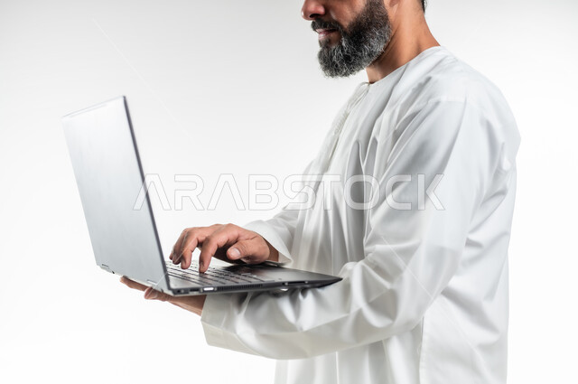 Using modern technologies and working remotely, diligence and completing the required tasks, following movies and series, browsing social networking sites, a close-up portrait from the side of an Arab Gulf Omani man wearing a dishdasha standing and holding a laptop in his hand, white background