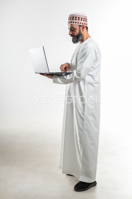 متابعة الأفلام والمسلسلات، تصفح مواقع التواصل الإجتماعي، الاجتهاد وانجاز الاعمال المطلوبة، بورتريه جانبي لرجل عربي خليجي عماني يرتدي الدشداشة والكمه يقف ويمسك بيده جهاز الحاسوب المحمول، استخدام التقنيات الحديثه والعمل عن بعد، الطول الكامل للجسم، خلفية بيضاء
