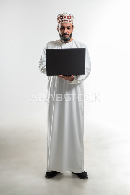 Watching movies and series, browsing social networking sites, the concept of diligence and completing the required work, a portrait of an Arab Gulf Omani man wearing a dishdasha and a kuma standing and holding a laptop in his hand, using modern technologies and working remotely, full body length, white background