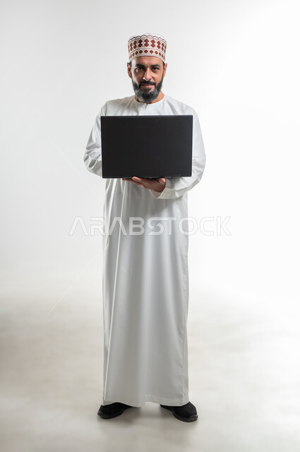 تصفح مواقع التواصل الإجتماعي، مفهوم الاجتهاد وانجاز الاعمال المطلوبة، بورتريه لرجل عربي خليجي عماني يرتدي الدشداشة والكمه يقف ويمسك بيده جهاز الحاسوب المحمول، مشاهدة الأفلام والمسلسلات، استخدام التقنيات الحديثه والعمل عن بعد، الطول الكامل للجسم، خلفية بيضاء
