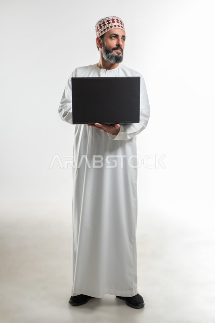 مفهوم الاجتهاد وانجاز الاعمال المطلوبة، بورتريه لرجل عربي خليجي عماني يرتدي الدشداشة والكمه يقف ويمسك بيده جهاز الحاسوب المحمول، تصفح مواقع التواصل الإجتماعي، مشاهدة الأفلام والمسلسلات، استخدام التقنيات الحديثه والعمل عن بعد، الطول الكامل للجسم، خلفية بيضاء