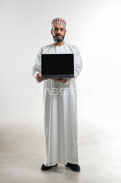 استخدام التكنولوجيا المتطورة للتسويق الرقمي الالكتروني، عرض تطبيقات وخدمات جهاز الحاسوب، بورتريه لرجل عربي خليجي عماني يرتدي الدشداشة و الكمه يحمل بيده جهاز الحاسوب المحمول بشاشة سوداء فارغة، الطول الكامل للجسم، خلفية بيضاء